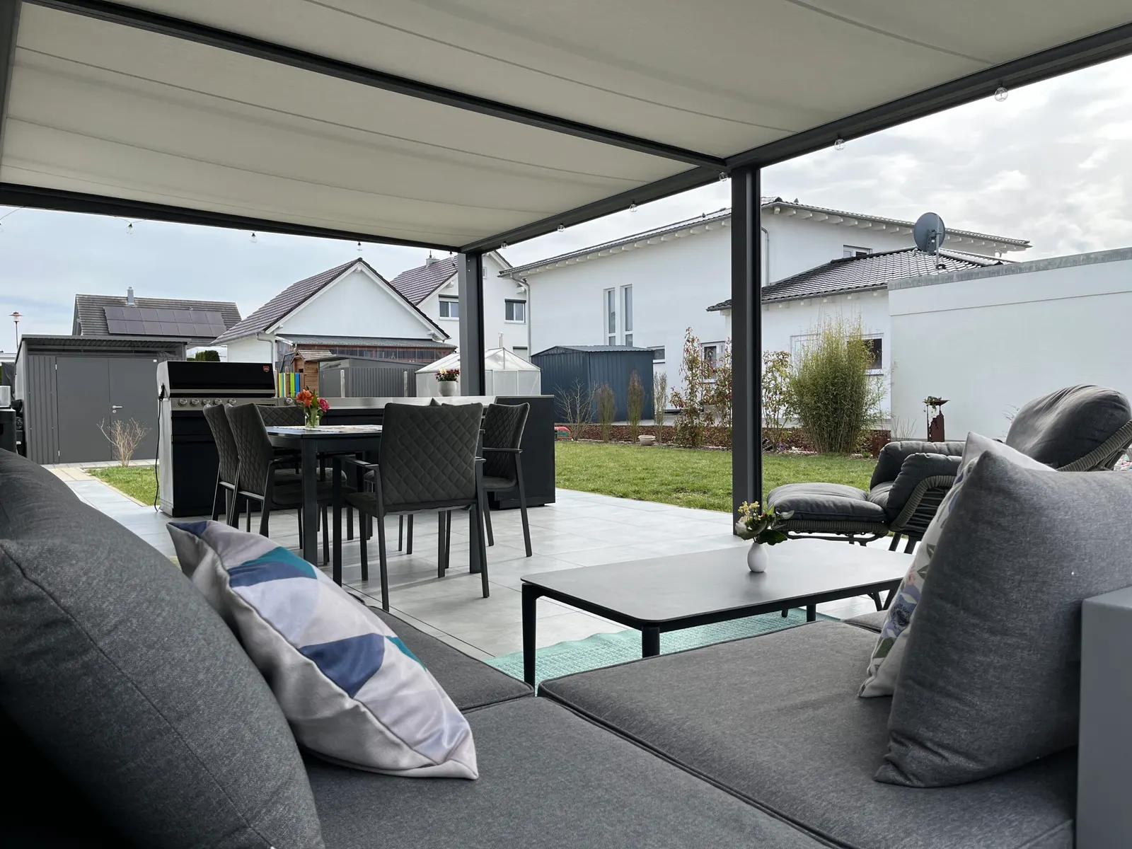 Blick aus der Lounge unter der ausgefahrenen Markise über Esstisch und Grill in den Garten des Neubaugebiets