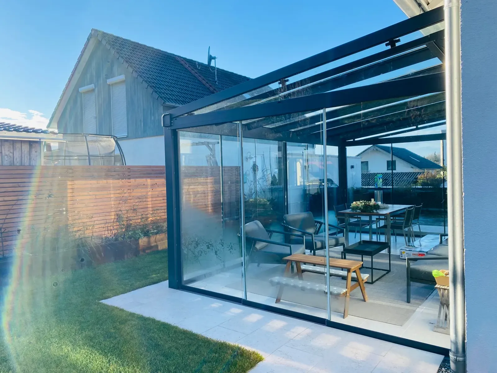 Blick an der Glasfront entlang auf Sitzgruppe und Holzbank unter dem Pultdach, hinter dem Lattenzaun steht ein Trampolin