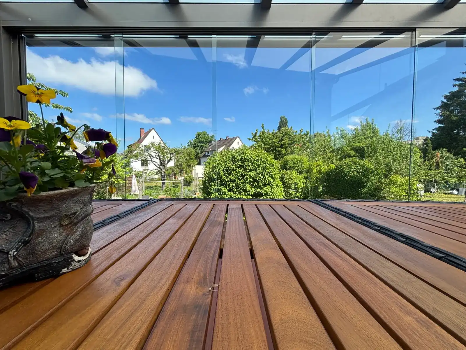 Blick vom Holzboden der Hochterrasse durch die rahmenlose Verglasung in die Nachbarschaft, links ein alter Pflanztopf mit Stiefmütterchen