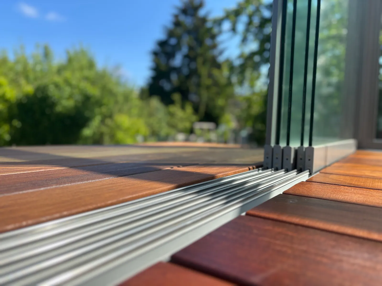 Nahaufnahme der Bodenschiene im Holzboden, rechts stehen die rahmenlosen Glasflügel geparkt hintereinander