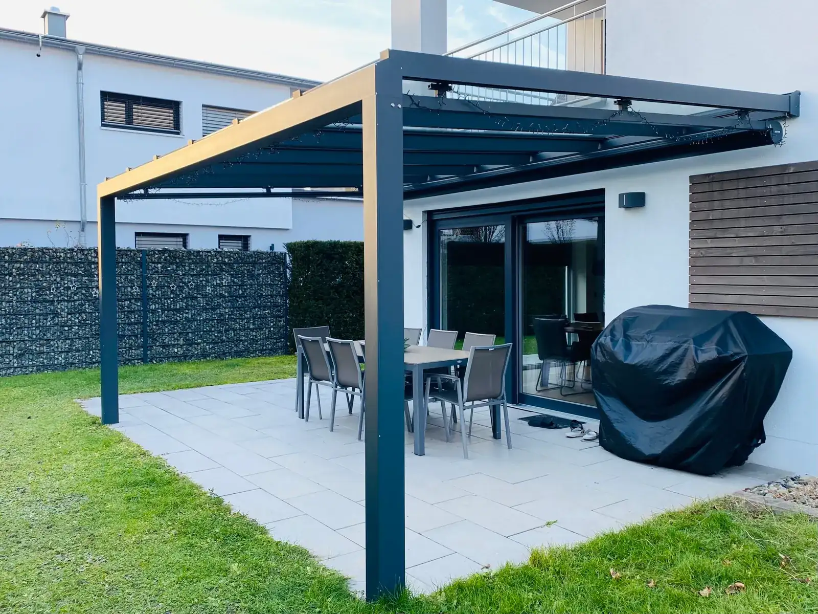 Das anthrazitfarbene Terrassendach von der Gartenecke aus, darunter der Esstisch mit Stühlen und ein abgedeckter Grill