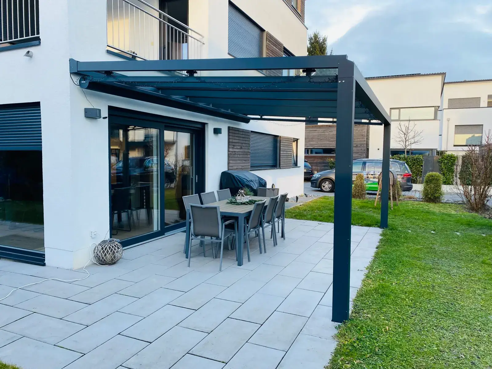 Blick über die hellen Terrassenplatten unter dem Glasdach, dahinter die Einfahrt mit Autos