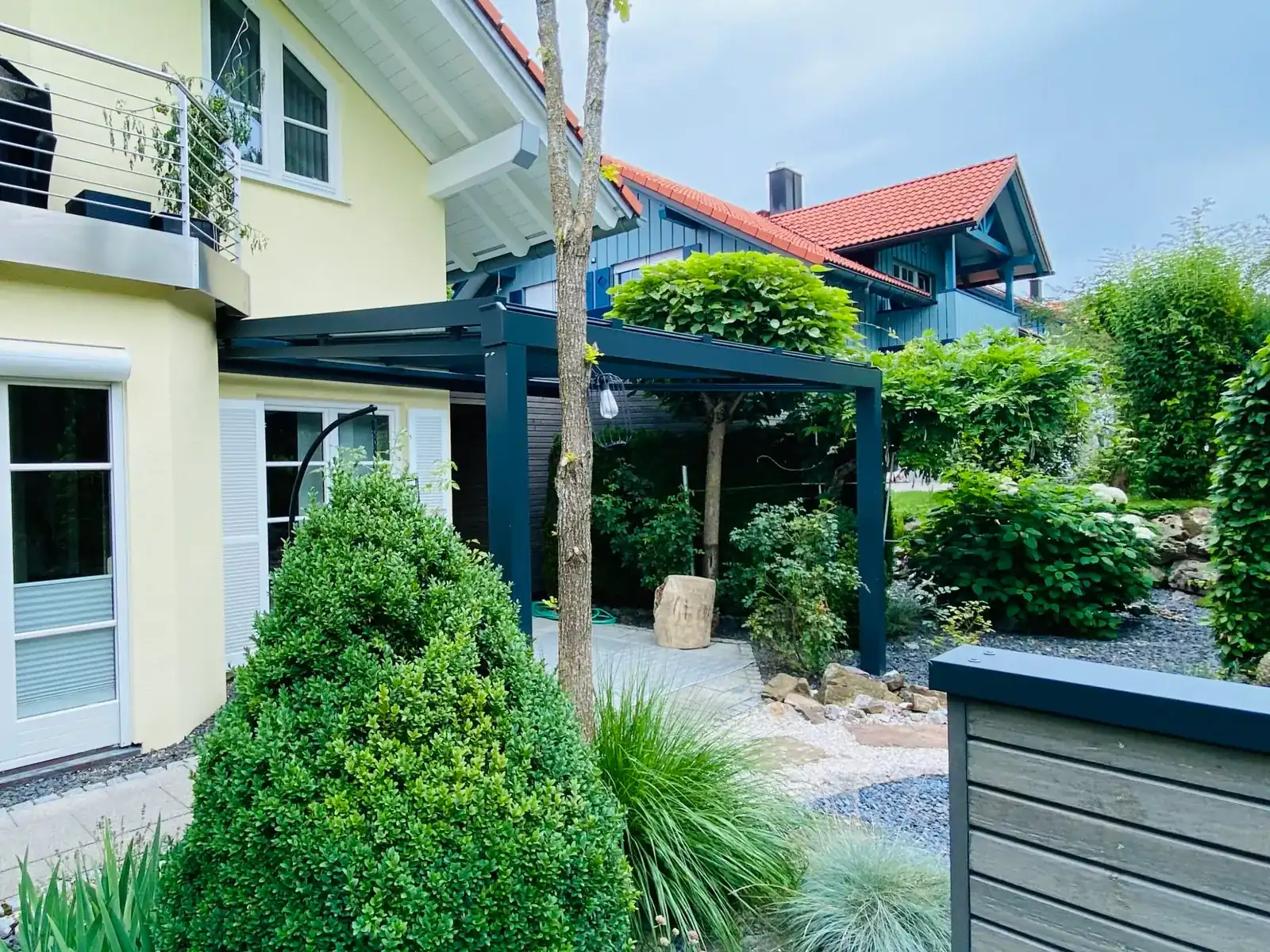 Das hellgelbe Wohnhaus in Weitnau: unter dem Balkon schließt das anthrazitfarbene Terrassendach an, davor Buchs und Gräser