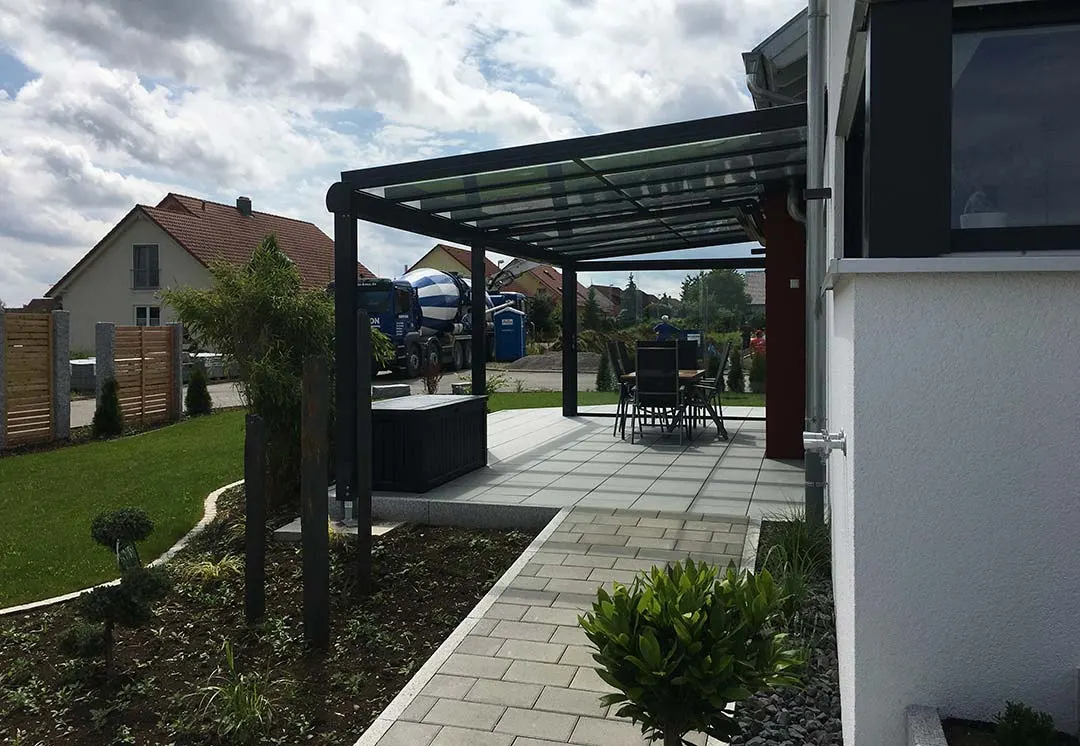 Blick unter das Glasdach auf anthrazitgrauen Stützen über die frisch gepflasterte Terrasse mit Sitzgruppe