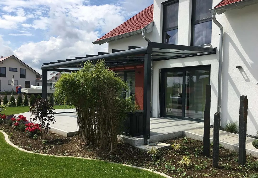 Die Überdachung von der Gartenseite: anthrazitgraues Pultdach auf schlanken Stützen, dahinter die dunkelrote Wandfläche und Bambus