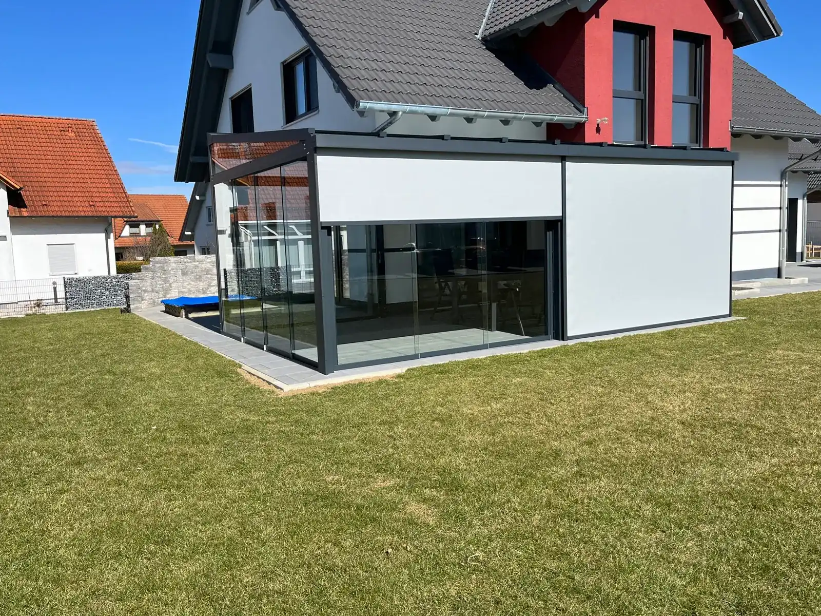 Gartenseite des Glasanbaus zwischen weißem und rotem Hausteil, die rechte Senkrechtmarkise ganz unten, die linke halb heruntergefahren