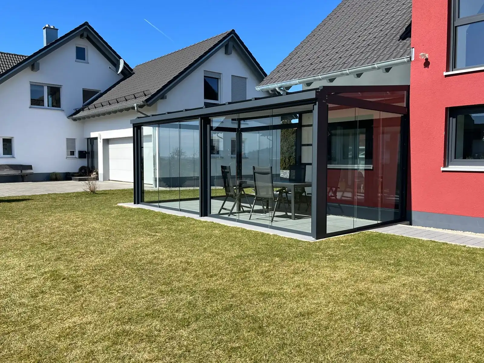 Der rundum verglaste Anbau läuft an der Ecke des roten Hausteils vorbei, innen stehen Gartentisch und Klappstühle