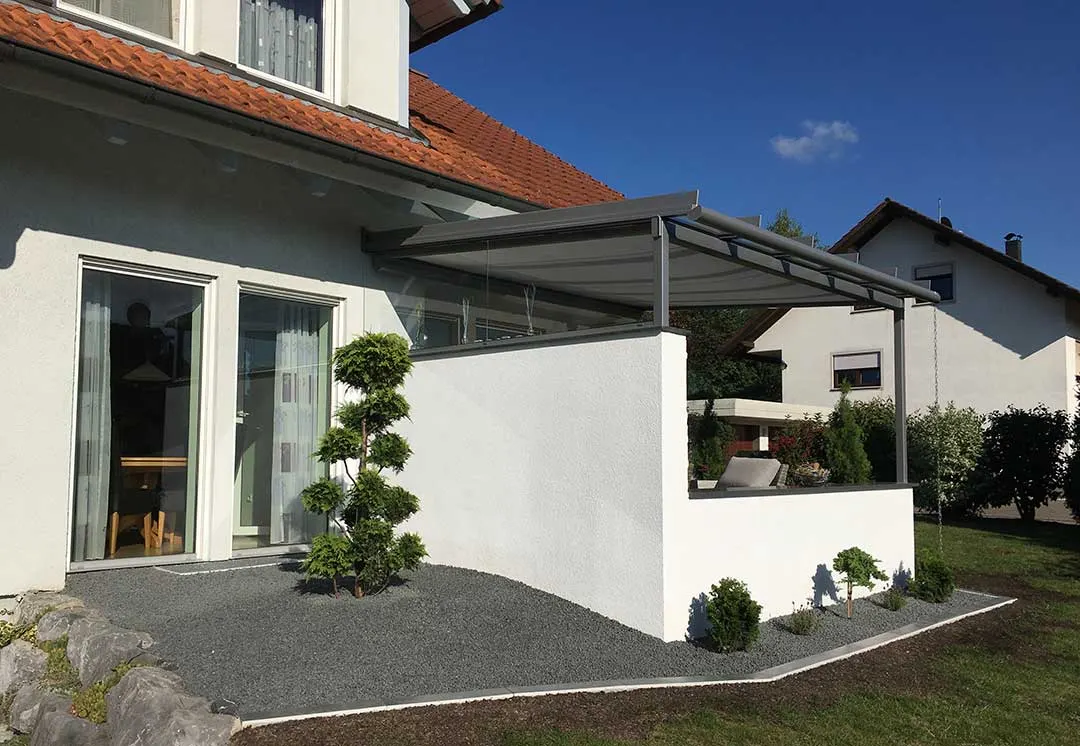 Die Terrassenüberdachung von außen: graues Pultdach über der weißen Brüstungsmauer, davor Splittbeete mit Formgehölzen