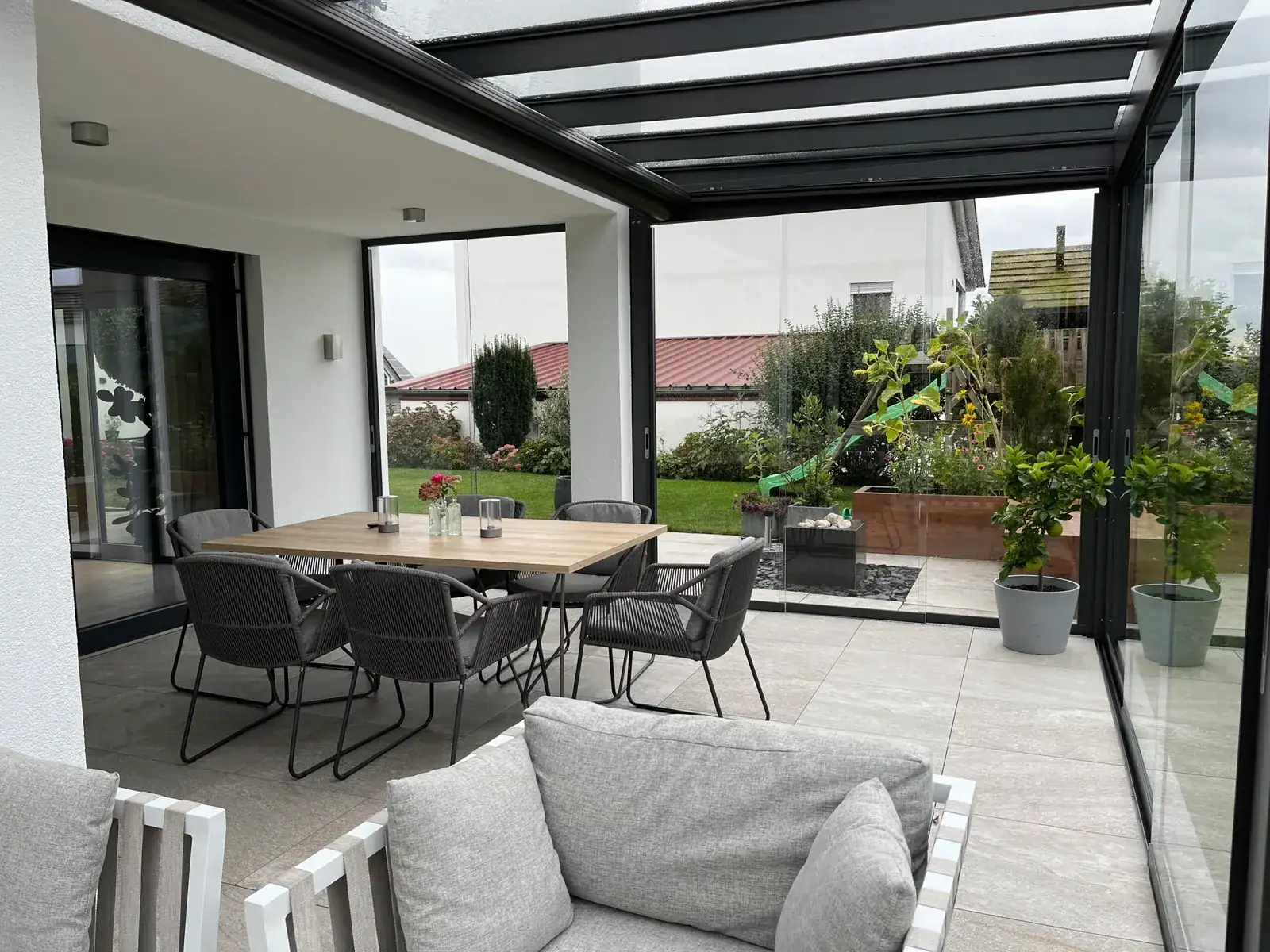 Esstisch und Lounge unter dem Glasdach, dahinter der Garten mit Spielturm