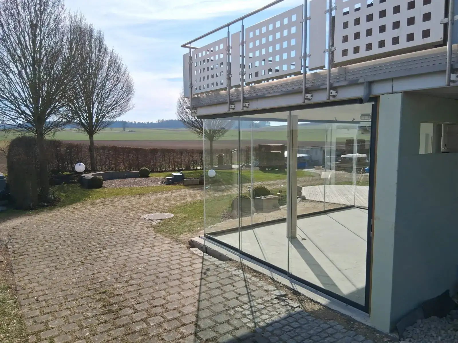 Die verglaste Ecke des Freisitzes unter dem Balkon, dahinter der Blick über die offenen Felder