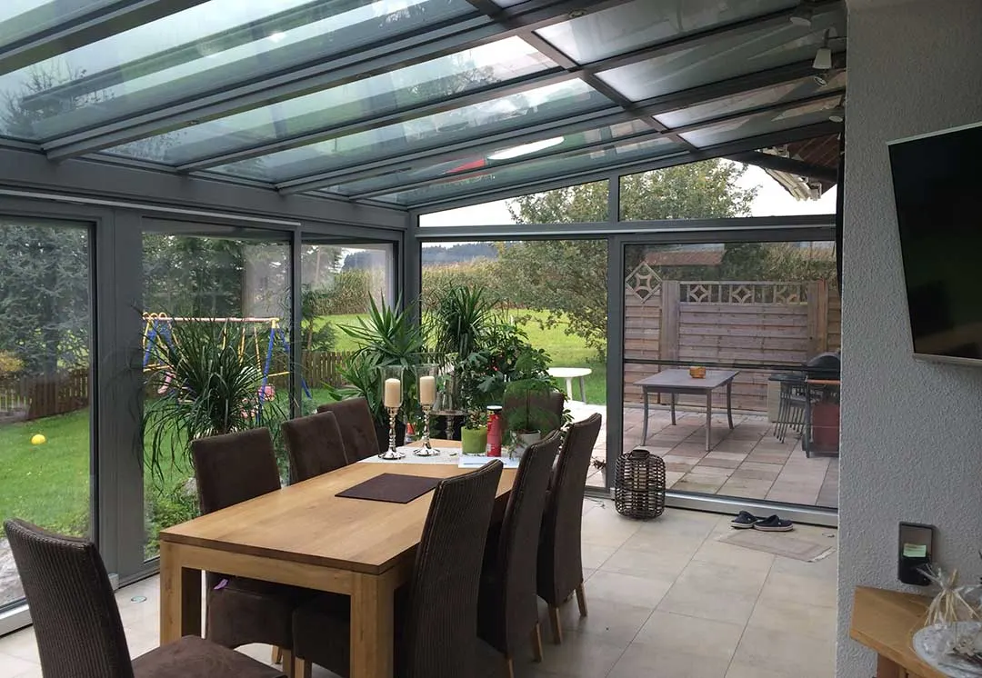 Der Wintergarten mit Blick zur gepflasterten Terrasse mit Holz-Sichtschutz und Grill