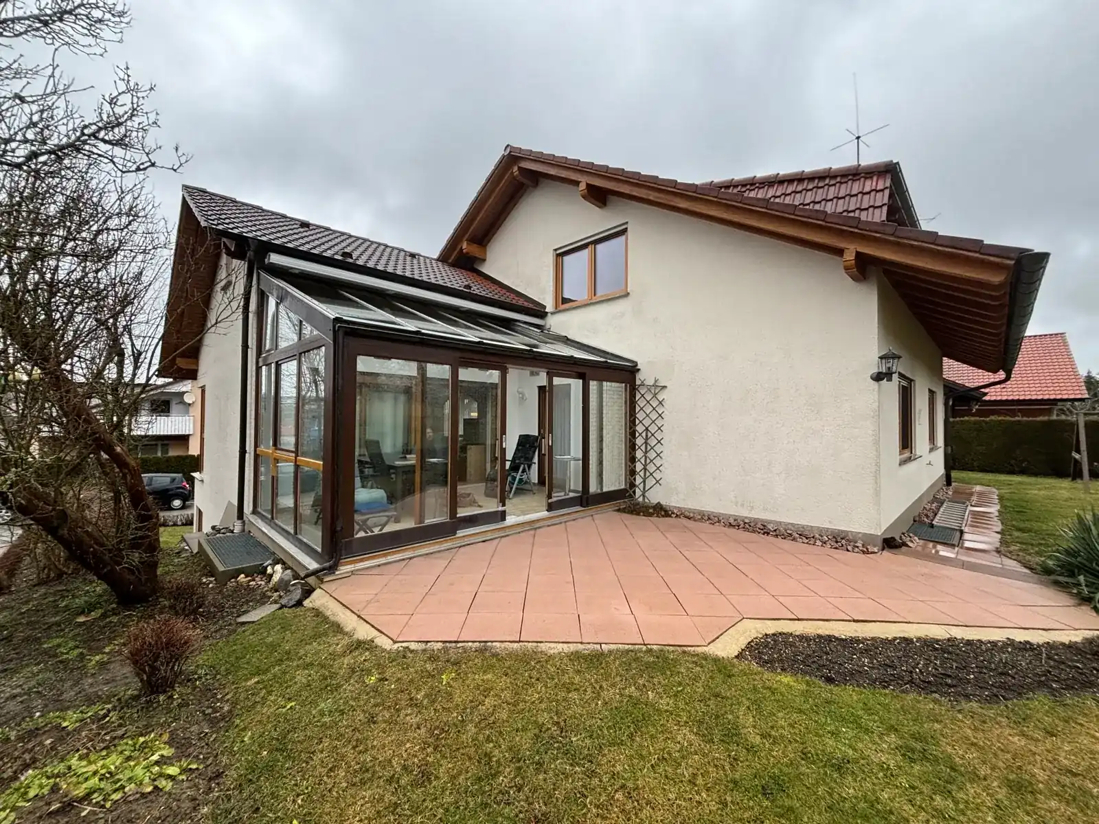 Der alte Holz-Wintergarten in der Hausecke an einem grauen Tag, davor die rötlichen Terrassenplatten