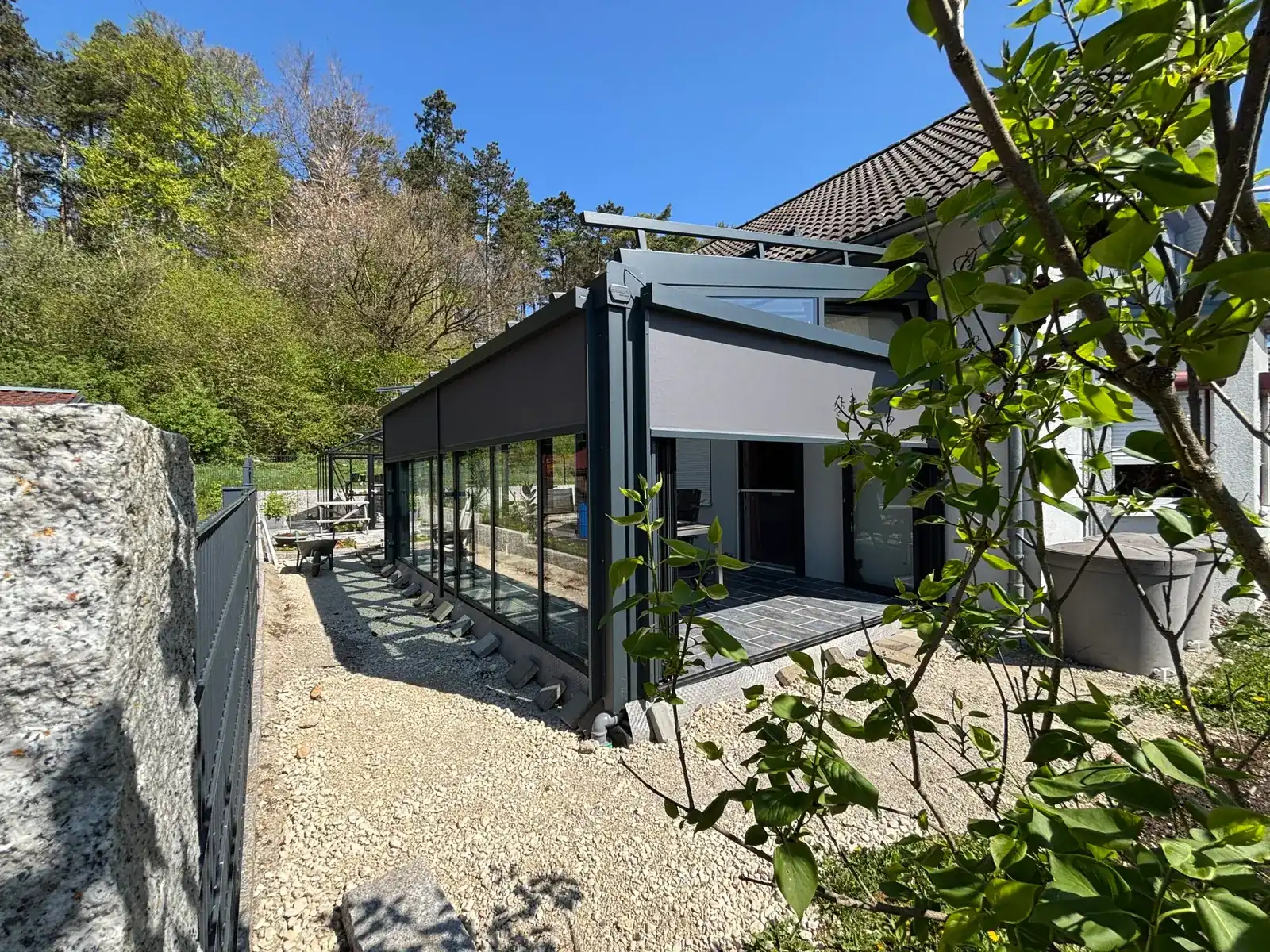 Der Wintergarten von außen: schmaler Anbau in Anthrazit am hellen Wohnhaus, an der Längsseite ist eine Senkrechtmarkise heruntergefahren, davor liegt noch Kies