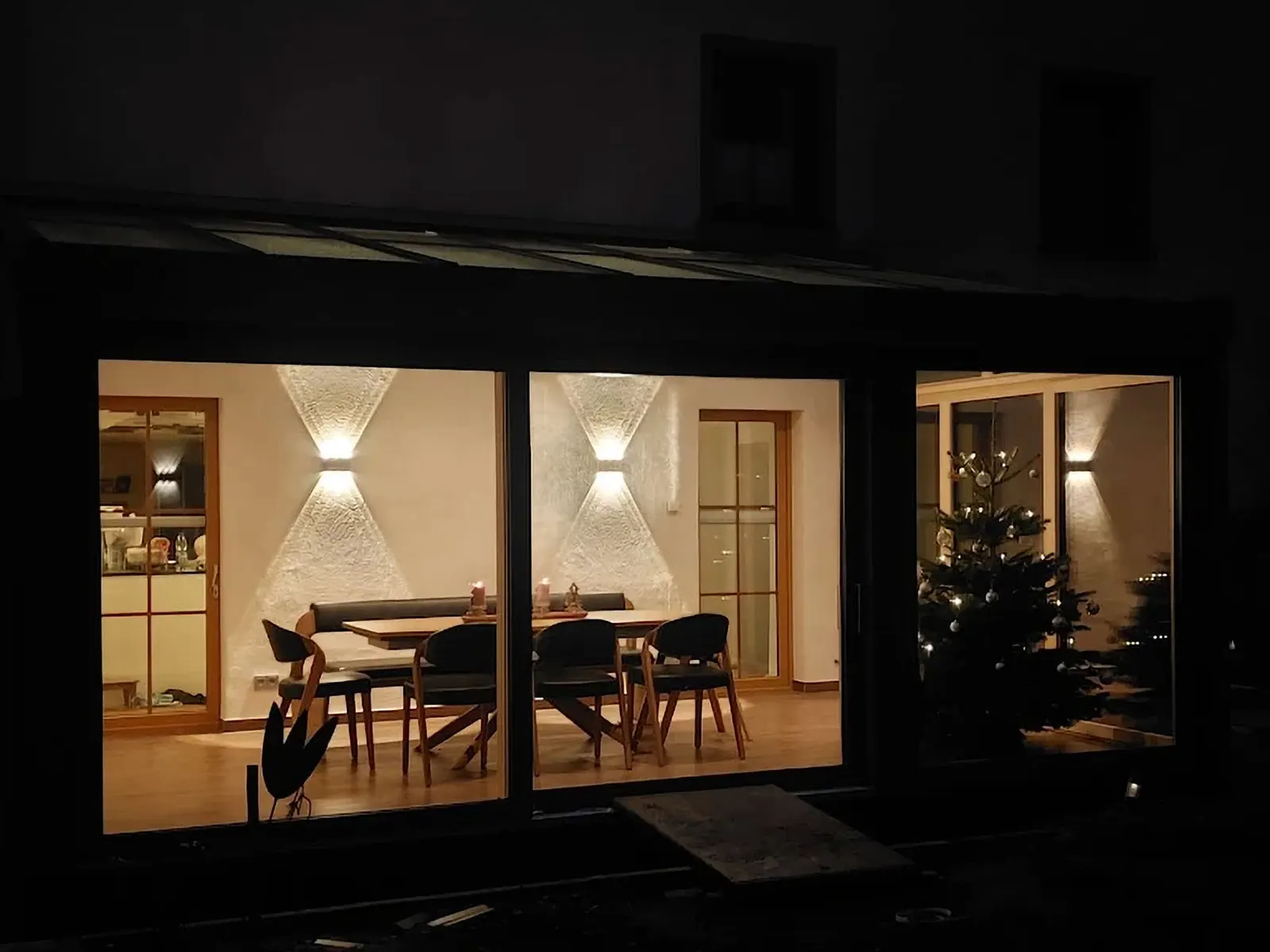 Blick von außen durch die Glasfront in den beleuchteten Wintergarten mit Esstisch, Eckbank und Christbaum