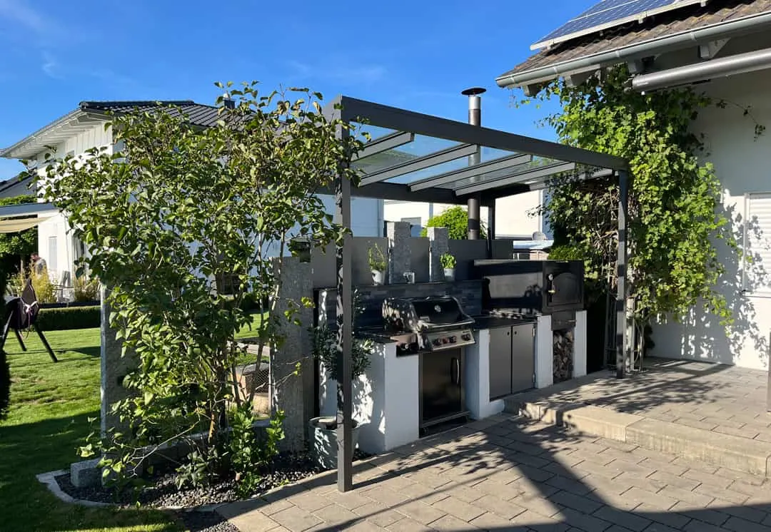 Das anthrazitfarbene Glasdach über der gemauerten Outdoor-Küche neben dem Haus, das Ofenrohr ragt über die Dachfläche hinaus