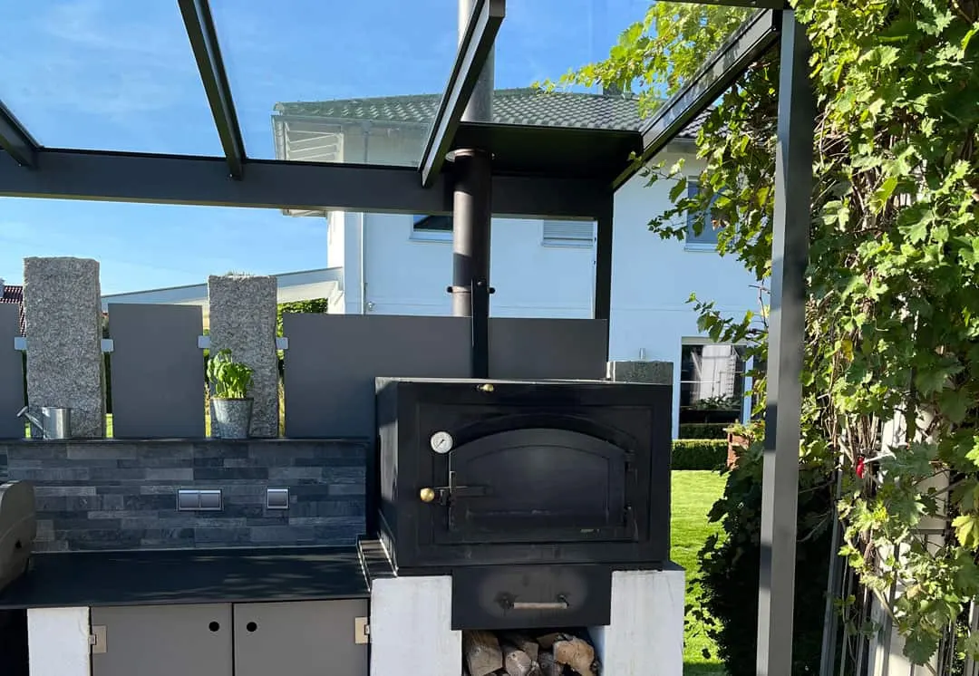 Der schwarze Holzbackofen unter dem Glasdach, sein Ofenrohr steigt durch die Dachebene, daneben ein geschlossenes Paneel statt Glas