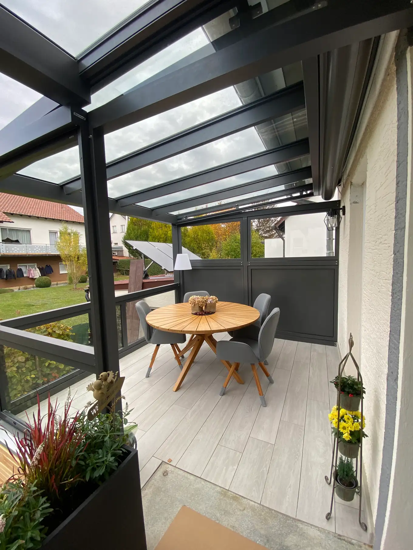 Der Sitzplatz unter dem Glasdach: rechts die Hauswand, hinten die Wand aus dunklen Paneelen mit dem Glasband darüber, zum Garten hin die niedrige Glasbrüstung