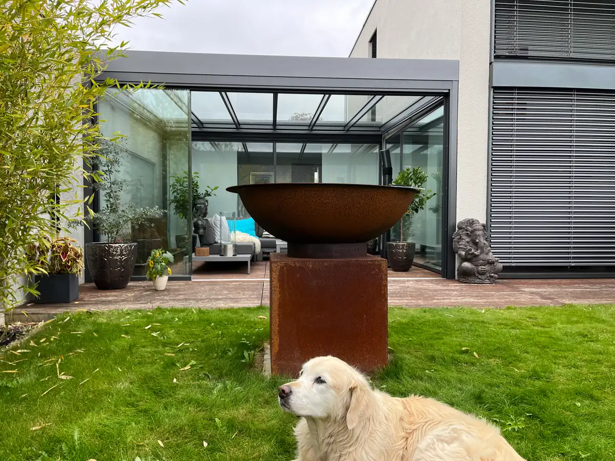 Blick über den Rasen auf das Glasdach in der Nische, vorn die Feuerschale aus Cortenstahl, der Hund liegt im Gras