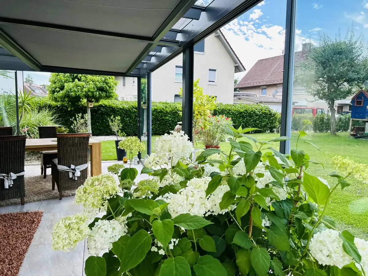 Die Unterglasmarkise von innen: das graue Tuch läuft unter dem Glasdach, dahinter der Blick in den Garten