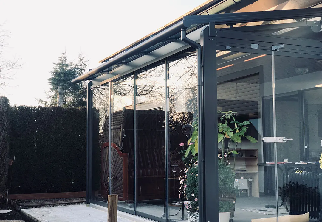 Das Glashaus von der Gartenseite: hinter der Glasfront der Strandkorb, darüber der Dachüberstand des Hauses, links die Hecke