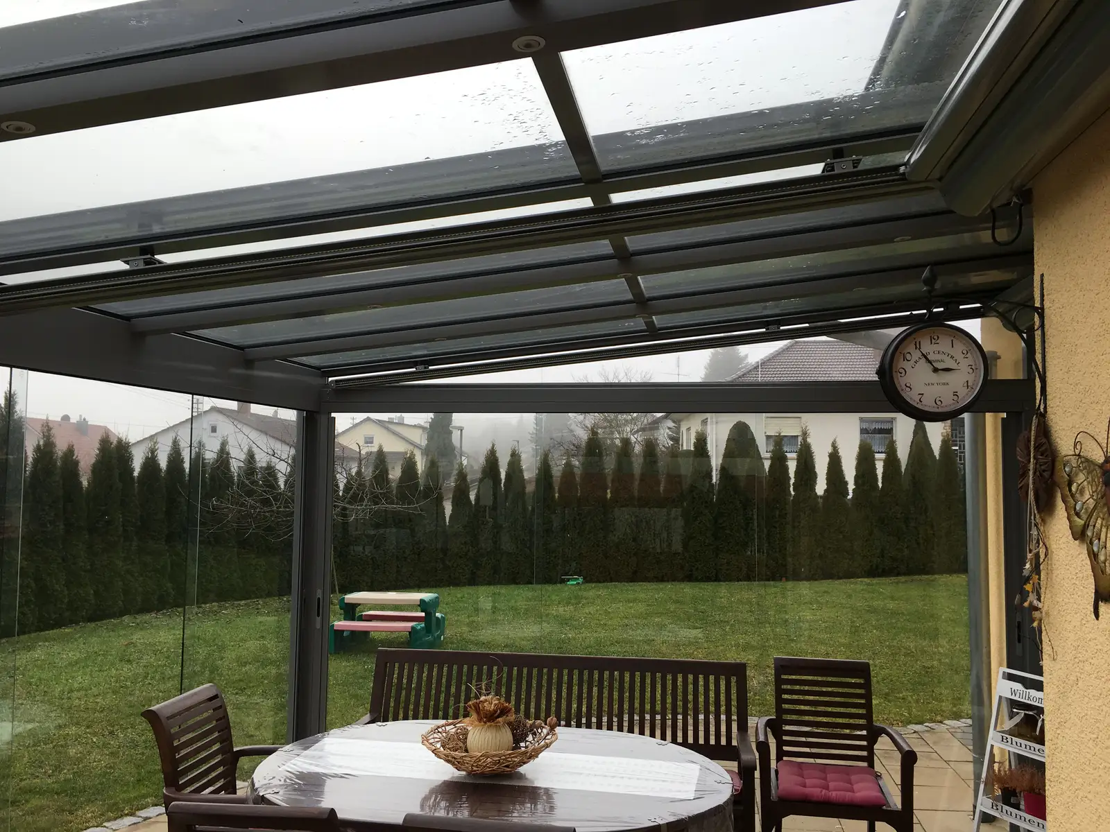 Blick vom Sitzplatz durch die Glasfront in den Garten, über dem Tisch die Sparren des Pultdachs, an der Hauswand eine Uhr