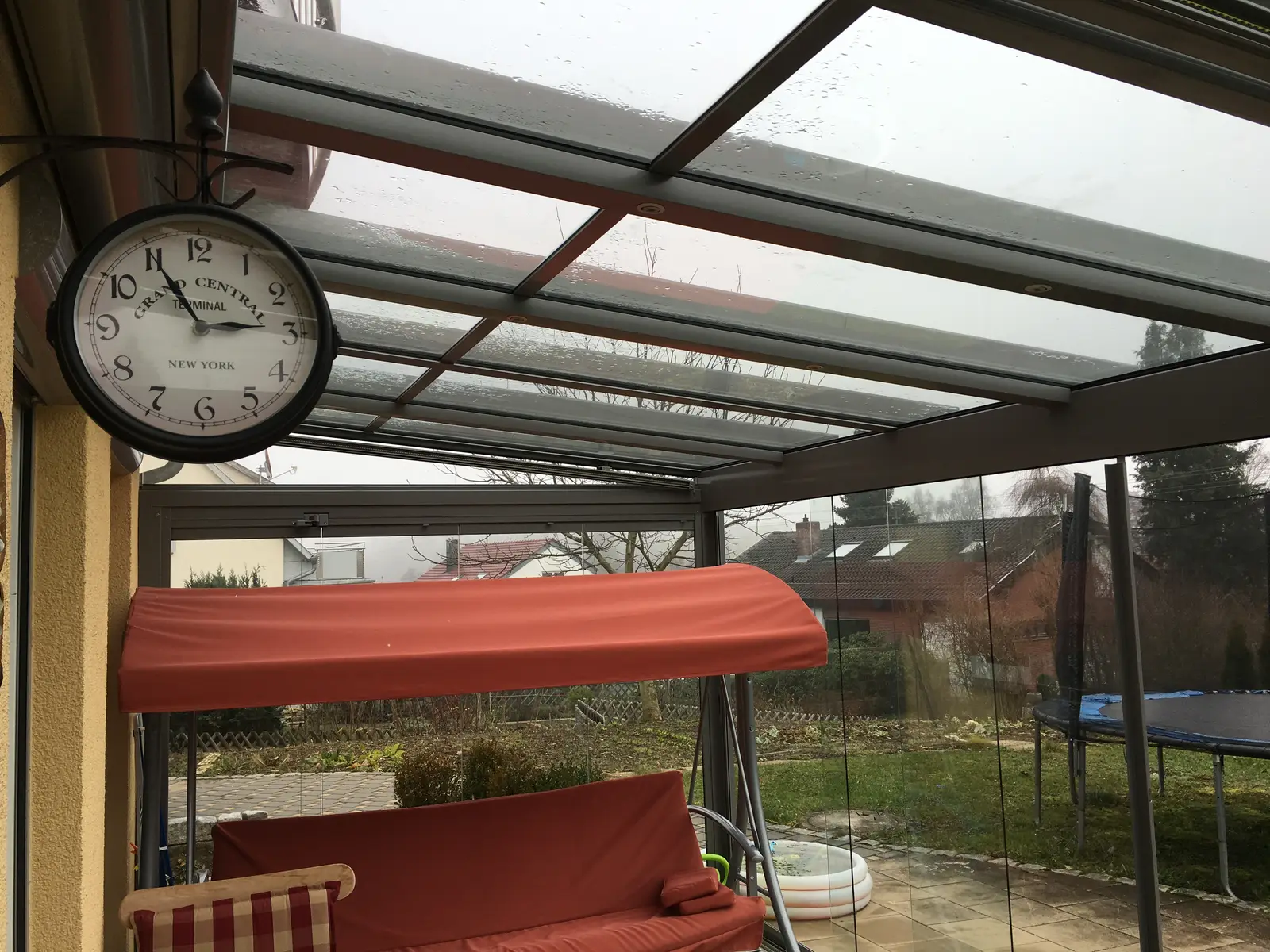Die Hollywoodschaukel unter dem Glasdach, dahinter durch die Scheiben das Trampolin im Garten
