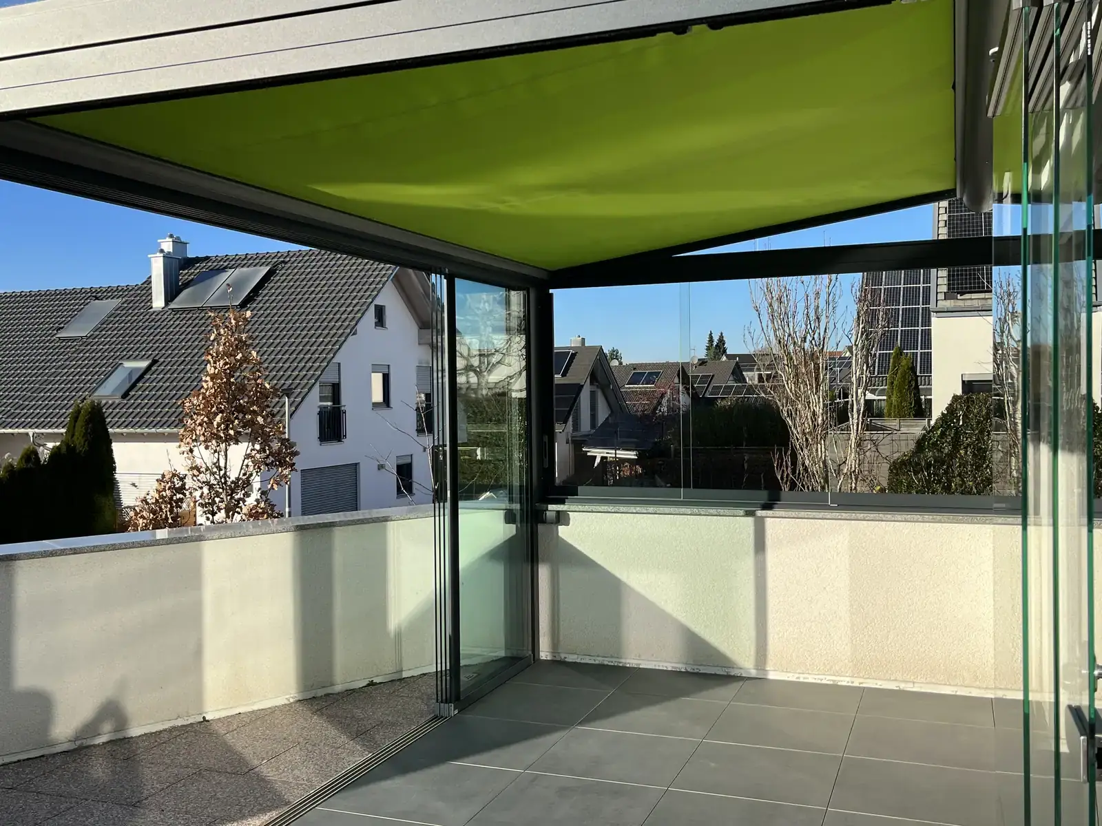 Die limonengrüne Unterglasmarkise unter dem Glasdach, dahinter die weiße Brüstung und die Dächer der Nachbarschaft