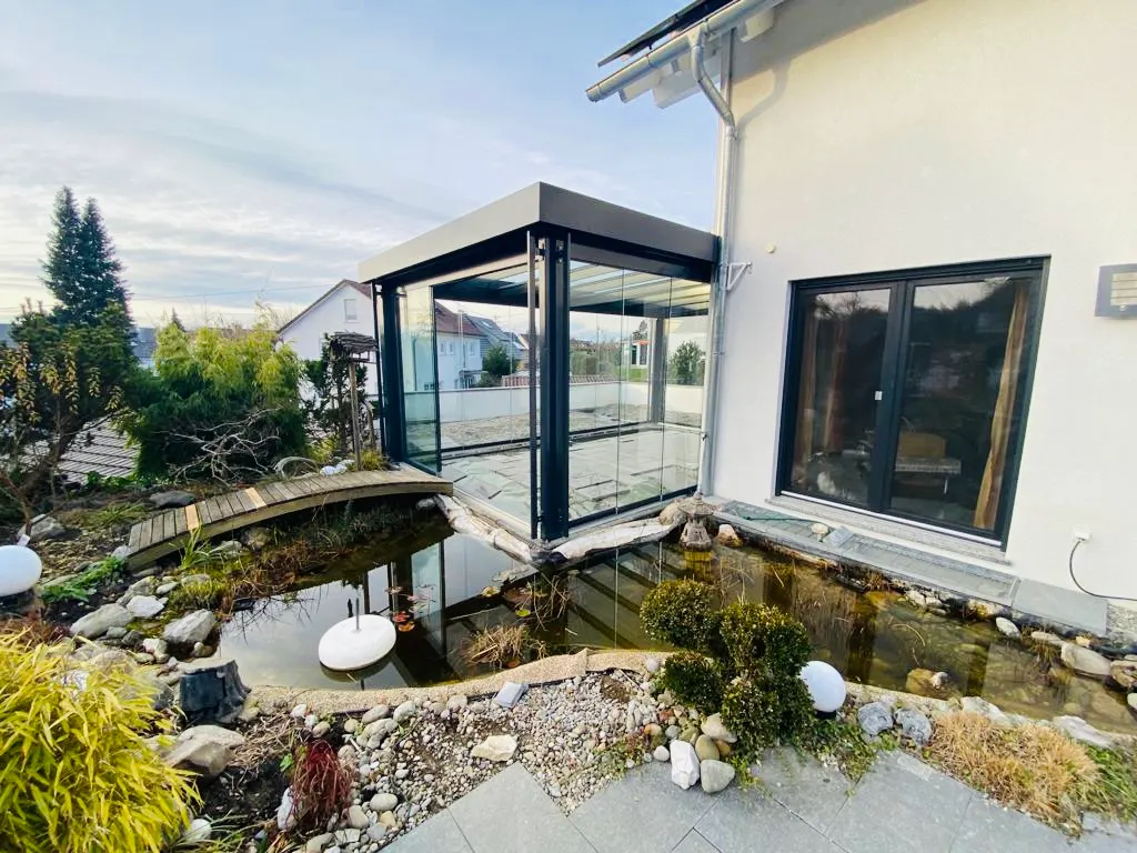 Das Glashaus steht direkt an der Kante des Gartenteichs, links führt ein Holzsteg über das Wasser zum Haus