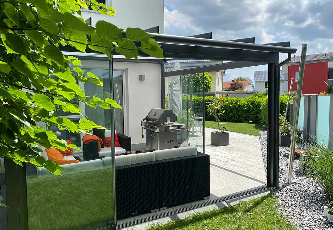 Blick durch Blätter auf das Pultdach: die Sparren stehen über den vorderen Träger hinaus, darunter Lounge und Grill