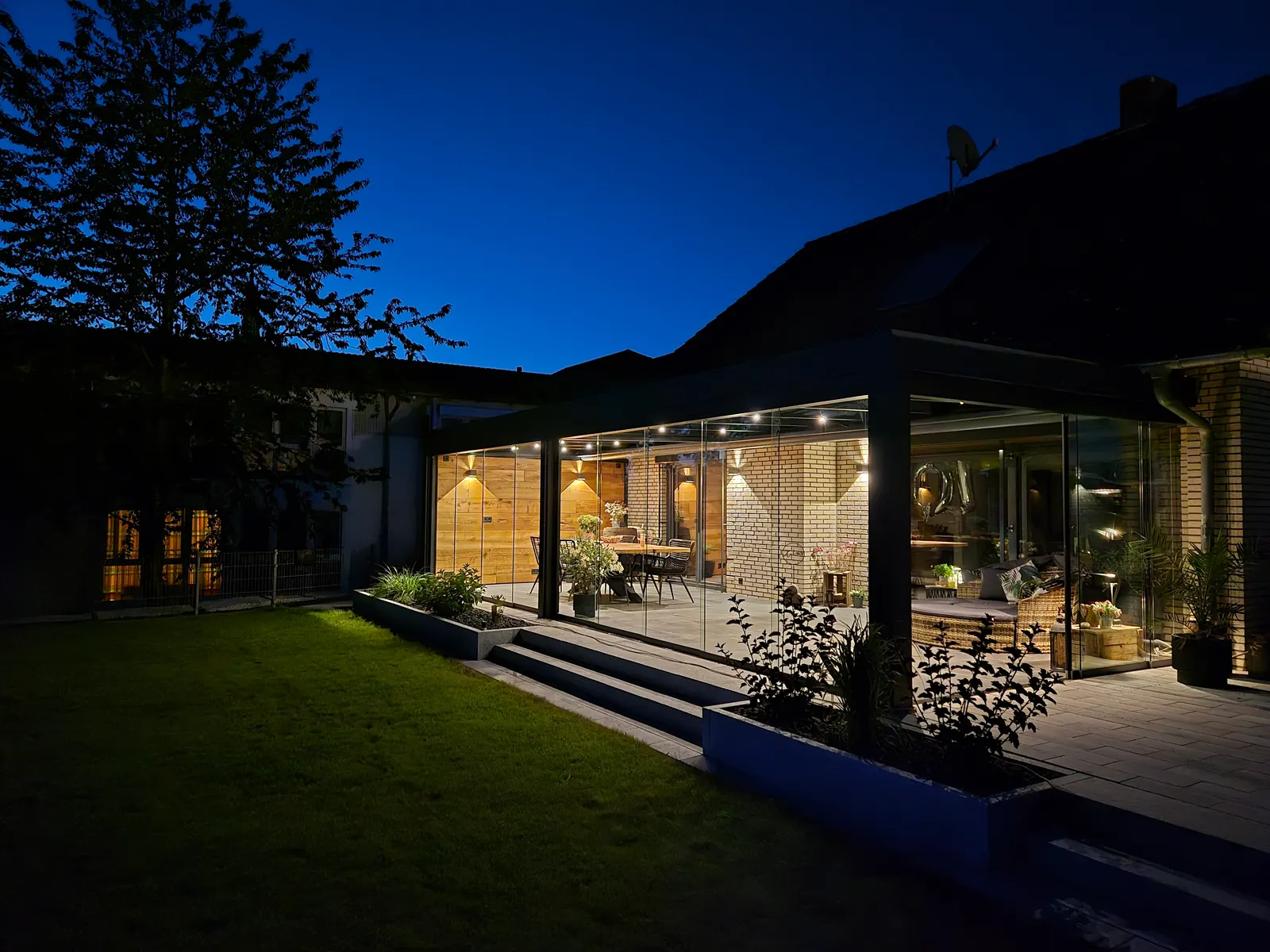 Das beleuchtete Glashaus am Abend von der Wiese aus, davor Granitstufen und bepflanzte Beete